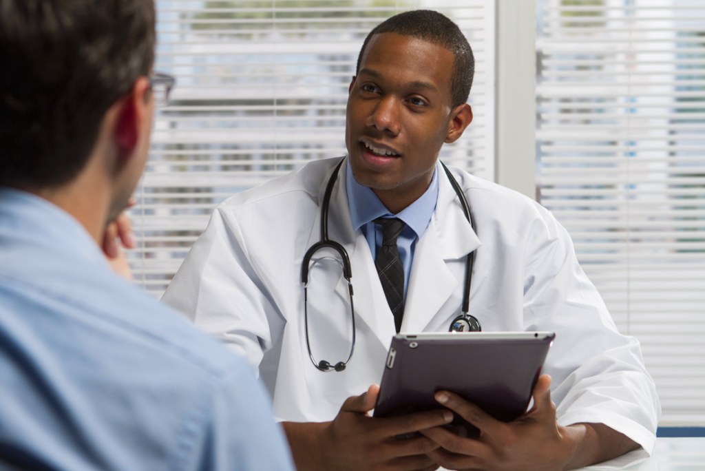 Imagem de um médico no consultório com um tablet nas mãos, atendendo um paciente.